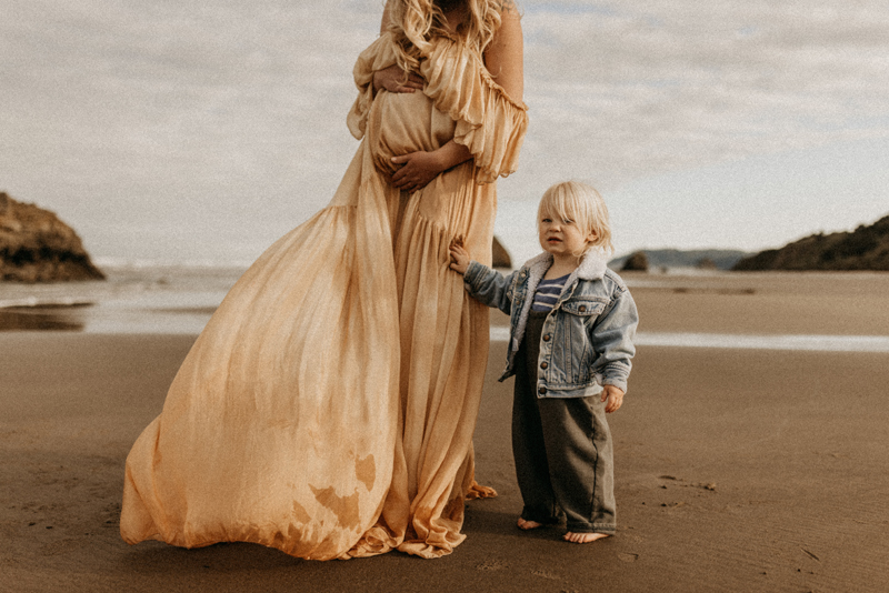 Maternity Photography, woman holding her belly on the beach with young toddler standing next to her